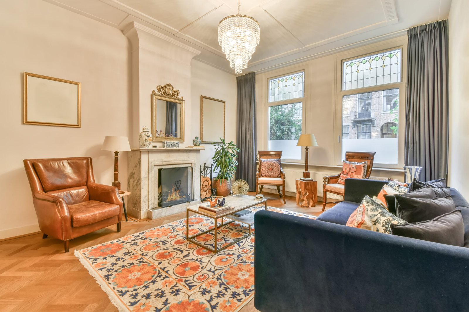 Elegant classic living room with high ceilings, stained glass windows, ornate white molding, marble fireplace, and eclectic decor elements including velvet sofa, floral rug, and leather armchair